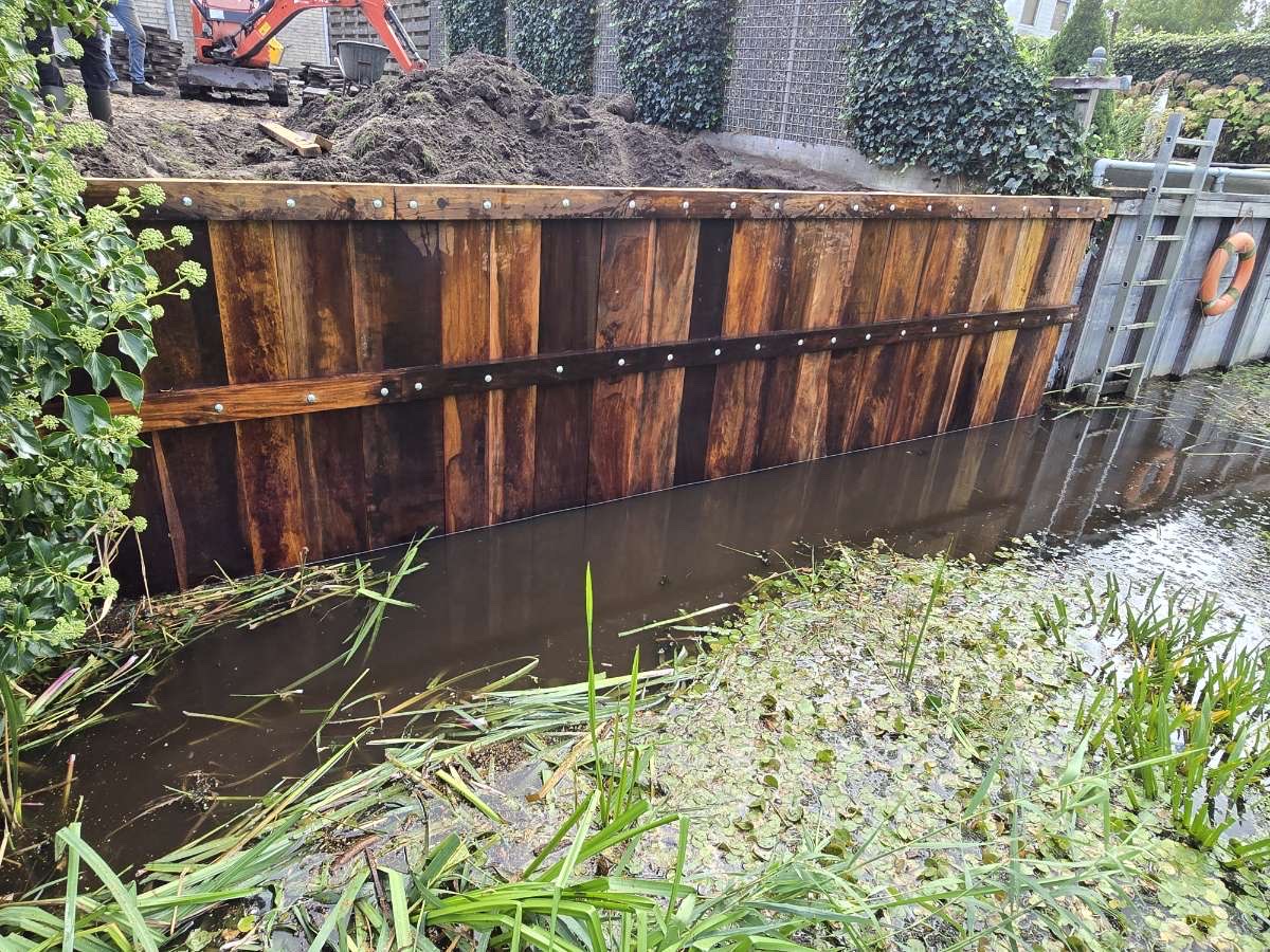 Net geplaatste hardhouten beschoeiing langs een slootkant met afgegraven grond en minigraver